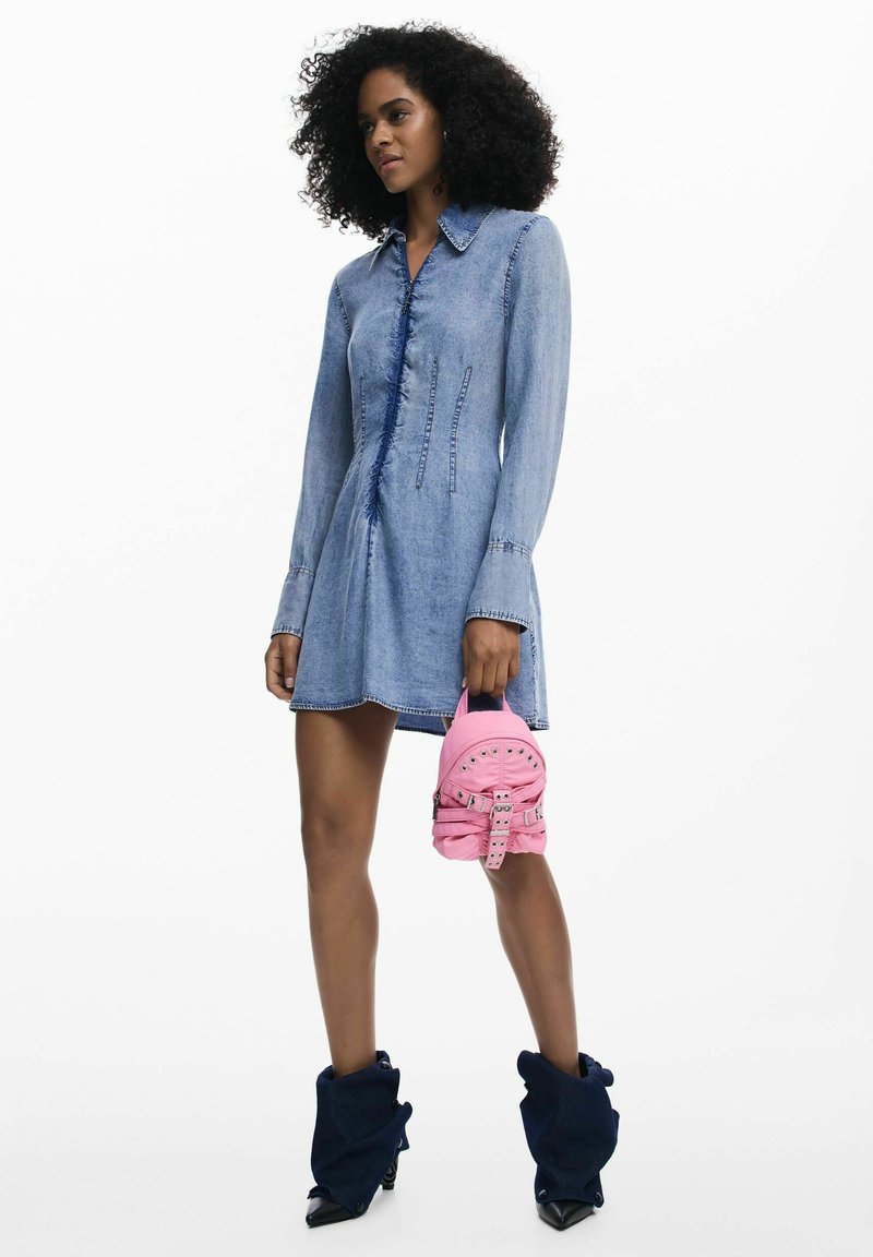 Kobieta w jasnoniebieskiej sukience dżinsowej z długimi rękawami i kołnierzykiem, trzymająca mały różowy plecak i nosząca ciemnoniebieskie, luźne botki.