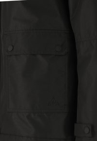 Zwarte jas met een grote voorzak, drukknopen en een gestructureerde afwerking. Beschikt over een subtiele logo in de onderste hoek.