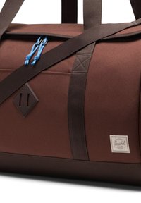 Sac de sport marron en tissu résistant avec des accents marron foncé, des cordons de tirage bleus, une patch logo en forme de diamant et des poignées robustes.