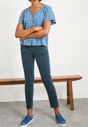 Haut bleu à manches courtes avec un motif floral, décolleté en V et détail de boutons, assorti à un pantalon slim en denim bleu foncé et des baskets bleues.