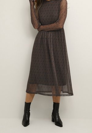 Robe midi marron à motif avec manches longues en voile, corsage ajusté et jupe fluide. Portée avec des bottines noires.