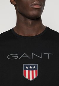 Černé tričko s krátkým rukávem s šedým logem "GANT" nad emblémem ve tvaru štítu, který je zdobený červenými, bílými a modrými pruhy a třemi bílými hvězdami.
