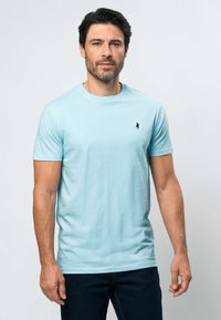 Vyras tamsiais plaukais ir barzda, dėvintis šviesiai mėlyną trumpomis rankovėmis marškinėlį ir tamsias kelnes, stovintis priešais vienspalvį šviesų foną.