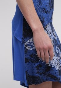 Main gauche reposant contre une jupe à motif paisley bleu et noir avec un haut bleu assorti.