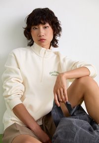 Sweatshirt zippé crème avec texte brodé vert, tissu doux, coupe décontractée, porté sur un short beige. Le modèle tient un tissu gris.