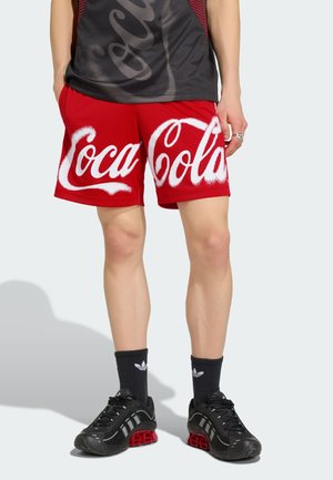 Persona que lleva pantalones cortos rojos con un gran texto blanco "Coca-Cola", calcetines negros Adidas, zapatillas deportivas negras y rojas, y una camisa negra y roja.