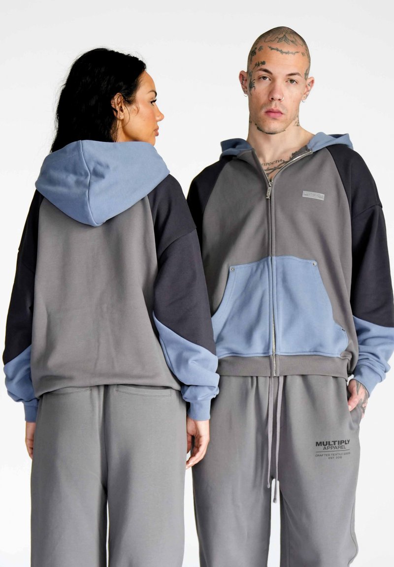 Šedá mikina s kapucňou na zips s modrými akcentmi, so predným vreckom, doplnená o zodpovedajúce šedé tepláky s detailom loga.