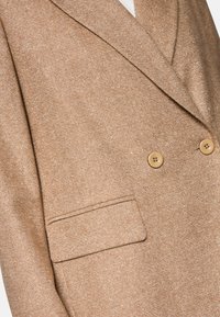 Hellbrauner strukturierter Blazer mit doppelter Knopfleiste, zwei runden Knöpfen und einer seitlichen Tasche.