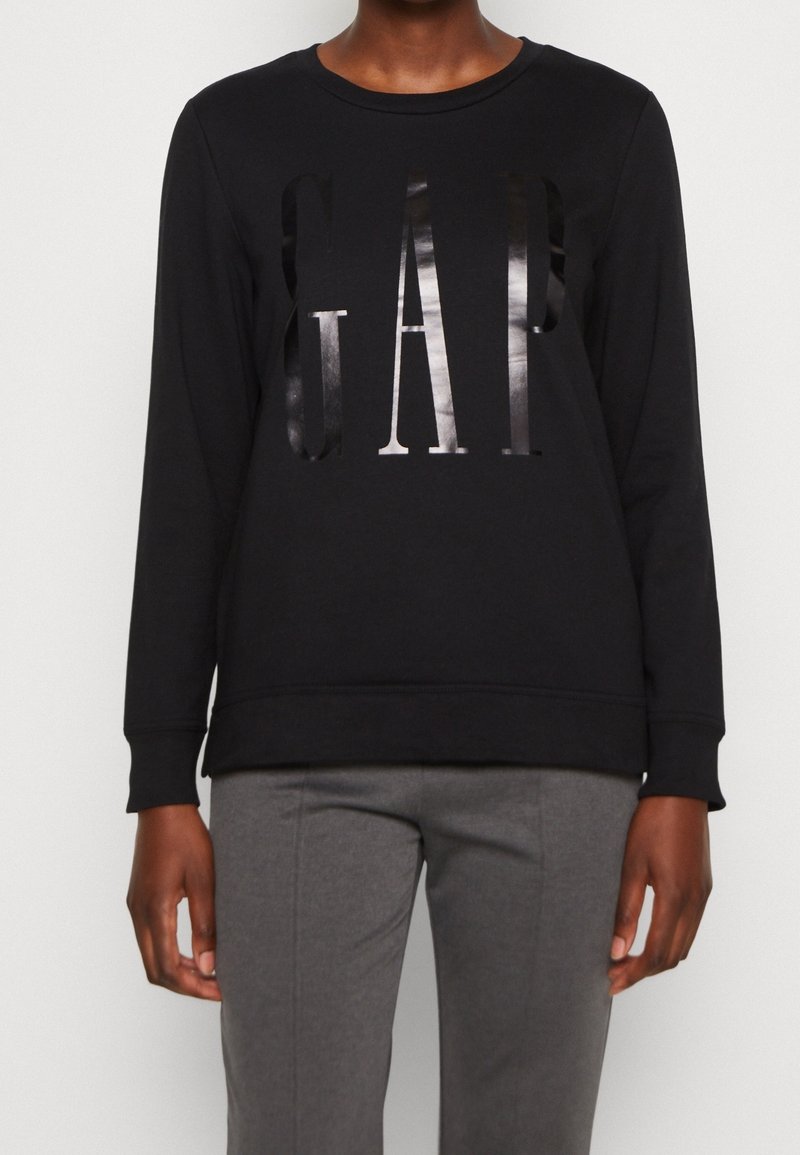Personne portant un t-shirt noir à manches longues avec des lettres brillantes « GAP » sur le devant et un pantalon gris, debout devant un fond uni.