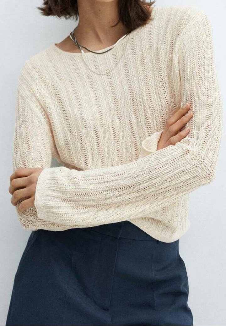 Vaaleanbeige neulottu neulepaita poimutetulla tekstuurilla, leveillä hihoilla ja väljällä istuvuudella, yhdistettynä tummiin räätälöityihin housuihin.