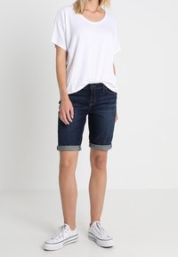 Bílá košile s krátkým rukávem, tmavě modré denimové bermudy s ohrnutými lemy, nošeny s bílými teniskami. Hladká tkanina a volný střih.