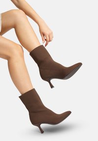 Botines marrones hasta el tobillo hechos de tela elástica, con punta afilada y tacón delgado. El diseño es ajustado y tiene una textura suave.