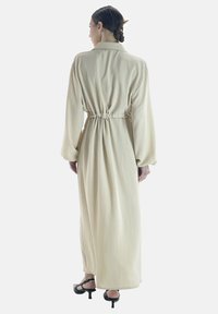 Robe longue beige avec des manches longues, taille cintrée et coupe décontractée. Texture douce avec une finition lisse et un col classique.