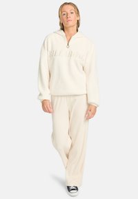 Crème-kleurige fleece trui met een halve rits en capuchon, met het "BILLABONG"-logo. Gecombineerd met lichtbeige geribbelde broek en zwarte sneakers.