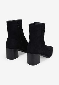Botas de ante negras a la altura del tobillo con punta redonda, cierre con cremallera y un tacón de bloque texturizado. El diseño presenta suaves pliegues en los lados.
