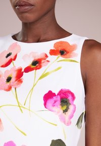 Blommig ärmlös topp med vit bakgrund, som har mönster av rosa, röda och gröna blommor. Slät tygkvalitet och rund halsringning.