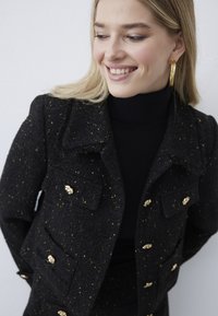 Usměvavá žena se světlými vlasy, která nosí velké zlaté kruhové náušnice a černou puntíkovanou bundu se zlatými knoflíky přes černý rolák.