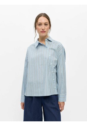 Femme aux cheveux attachés portant une chemise boutonnée à rayures verticales bleues et blanches et un pantalon large bleu foncé.