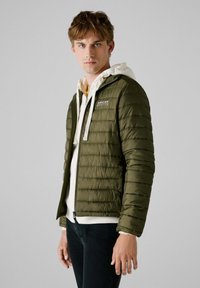 Chaqueta aislante verde oliva con diseño de acolchado horizontal, cierre de cremallera y cordones blancos. Llevada sobre una sudadera ligera y jeans.