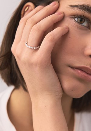 Young woman with short brown hair touching her face, wearing a silver ring with multiple clear gemstones on her ring finger.