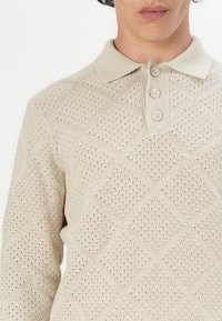 Polo de punto beige con un patrón texturizado en forma de diamante, que presenta un cuello y una tapeta de tres botones, confeccionado en una mezcla de algodón transpirable.