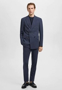 Costume bleu foncé à double boutonnage avec deux rangées de boutons noirs, pantalon slim fit et chaussures de ville noires. Tissu lisse, design ajusté.