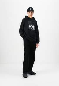 Mladý muž stojí oblečený v čiernej mikine Helly Hansen, čiernych nohaviciach, čiernej čiapke s bielym logom a čiernych topánkach na bielom pozadí.