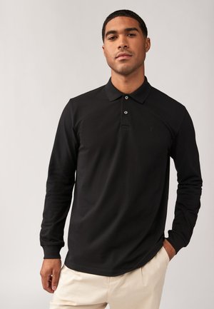 Hombre con una camisa de polo negra de manga larga y pantalones beige, de pie frente a un fondo claro y liso con una mano en el bolsillo.