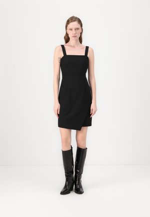 Robe noire sans manches avec un décolleté carré et un ourlet asymétrique, associée à des bottes en cuir noir gaufré crocodile arrivant au genou.