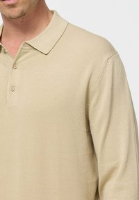 Tricou polo bej cu mânecă lungă, cu o textură canelată, guler clasic și închidere cu trei butoane, evidențiind linii curate și o croială ajustată.