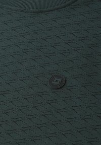 Tissu vert foncé avec un motif abstrait de lignes noires répétées et un petit emblème rond noir comportant un design géométrique près du centre.