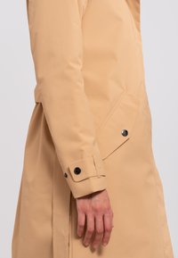 Vedenkestävä beige takki sileällä tekstuurilla, jossa on sivutasku ja mustat napit hihassa ja taskun läpällä.