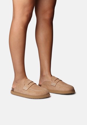 Beżowe zamszowe buty wsuwane z zaokrąglonym noskiem, z białym przeszyciem i ozdobnym paskiem na cholewce. Płaska podeszwa dla codziennego komfortu.