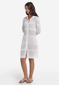 Biała sukienka na szydełku z zapięciem na guziki, z wzorami poziomymi i diamentowymi, długimi rękawami oraz długością do kolan.
