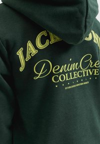Sweat à capuche vert foncé avec un texte jaune indiquant "Denim Crew Collective Worldwide, Established Nineteen Ninety" et de grandes lettres partiellement visibles au-dessus.