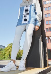 Jeansjacke mit grauem Hoodie, versehen mit schwarz-weißen Streifen, kombiniert mit weißen Leggings und weißen Sneakers. Texturiertes und lässiges Design.