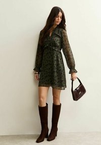 Vestido verde oliva con estampado floral, mangas largas transparentes y detalles con volantes. Combinado con botas marrones hasta la rodilla y un bolso marrón.