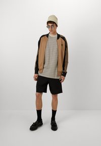 Ruskea ja musta vetoketjullinen urheilutakki, harmaa raidallinen t-paita, mustat shortsit, mustat sukat, mustat kengät ja beige neulottu pipo.