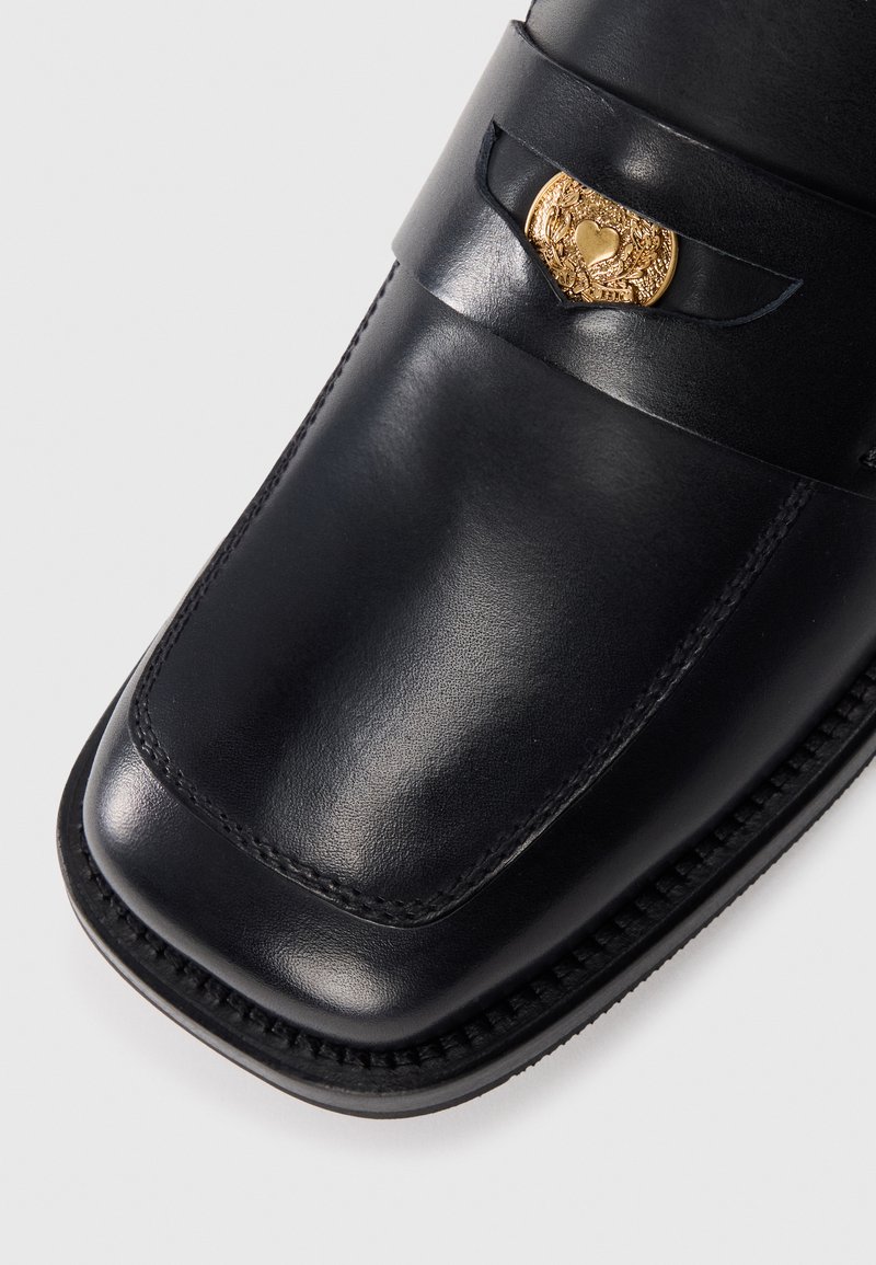 Zwarte leren instapper met een gladde afwerking, ronde neus en decoratieve gouden hardware-accent op een band. Gestikte details langs de rand.