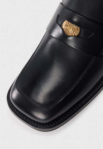 Sapatos loafers de couro preto com bico redondo, com detalhes decorativos em dourado e costura ao longo da borda. Textura suave.