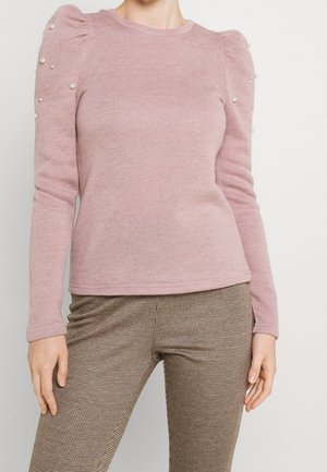 Femme portant un pull rose poudré à manches longues avec des épaules bouffantes et des ornements en perles, associé à un pantalon marron à carreaux.