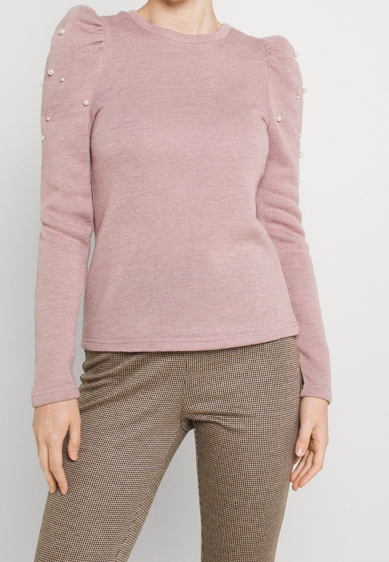 Femme portant un pull rose poudré à manches longues avec des épaules bouffantes et des ornements en perles, associé à un pantalon marron à carreaux.