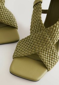 Sandale cu toc înalt din piele verde măsliniu, cu curele împletite, vârf pătrat și suprafață texturată. Toc subțire și detalii cu finisaj delicat.
