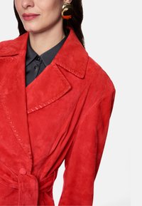 Red suede jacket with a large collar, visible stitching details, and a tie belt. Worn over a black shirt; features a smooth texture and pronounced shape.