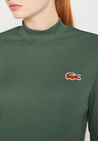 Zelený rebrovaný rolák s malým oranžovým logom krokodíla na hrudi. Má textúrovanú látku a priliehavý strih.