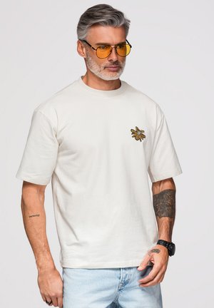 Mand med gråt hår og skæg iført gule brilleglas, hvid t-shirt med bi-patch, lyse jeans og tatoveringer på begge arme står mod hvid baggrund.