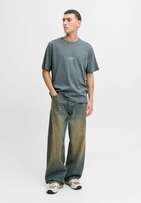 Graues Rundhals-T-Shirt mit kleinem Logo, kombiniert mit weit geschnittenen, ausgebleichten blauen Jeans und beigen Sneakers, vor einem schlichten Hintergrund.