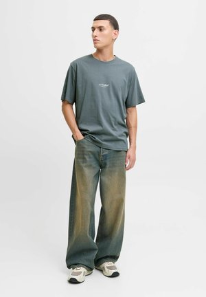 T-shirt gris à col rond avec un petit logo, associé à un jean bleu délavé à jambe large et des baskets beiges, sur fond uni.