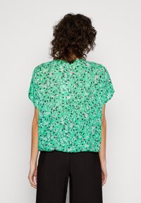 Blusa floral verde con mangas cortas, de tela ligera y corte holgado. Presenta un patrón diverso de flores en blanco y negro sobre un fondo verde.