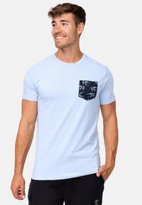 Camiseta de algodón azul claro con cuello redondo y mangas cortas, que presenta un bolsillo de parche en azul marino con un patrón de pájaros en blanco.